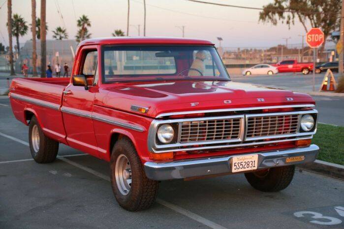 Ein roter Oldtimer pickup aus 1970 der Marke Ford auf einem Parkplatz. Im Hintergrund Palmen und Straße.