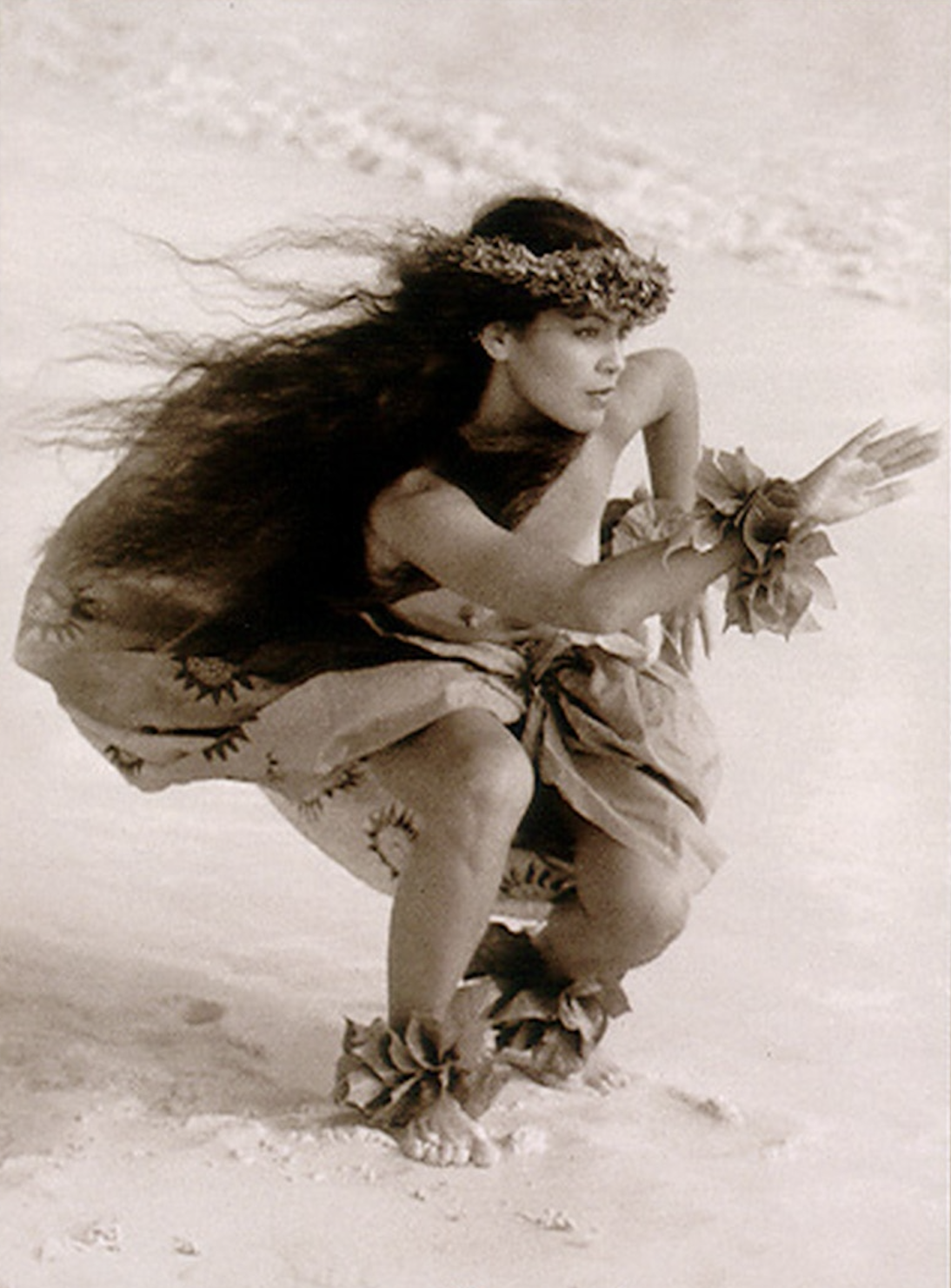 Eine Schwarz-Weiß Fotografie mit leichtem Sepia-Farbstich. Zu sehen ist eine junge Hula Tänzerin am Strand, während eines Tanzes.