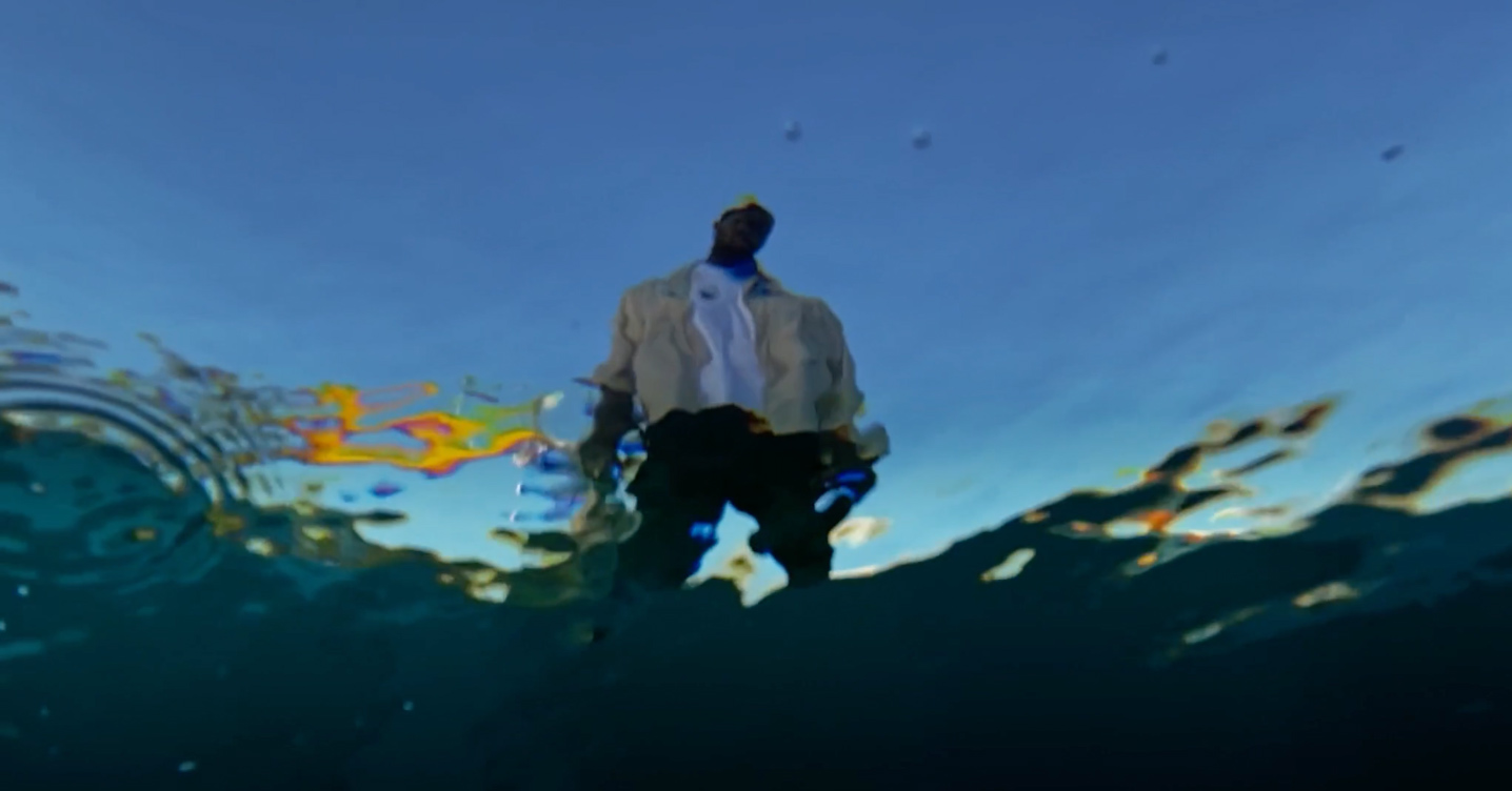 Ein Mann steht am Rand eines Schwimmbeckens. Der Blick geht von der Wasserunterfläche auf ihn hinauf. Die Silhouette des Mannes ist verschwommen.